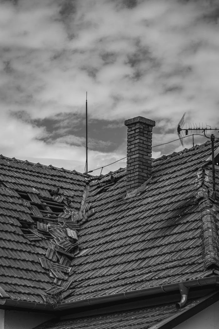 services-02 A grayscale photo of a broken roof with a chimney and antenna under a cloudy sky.